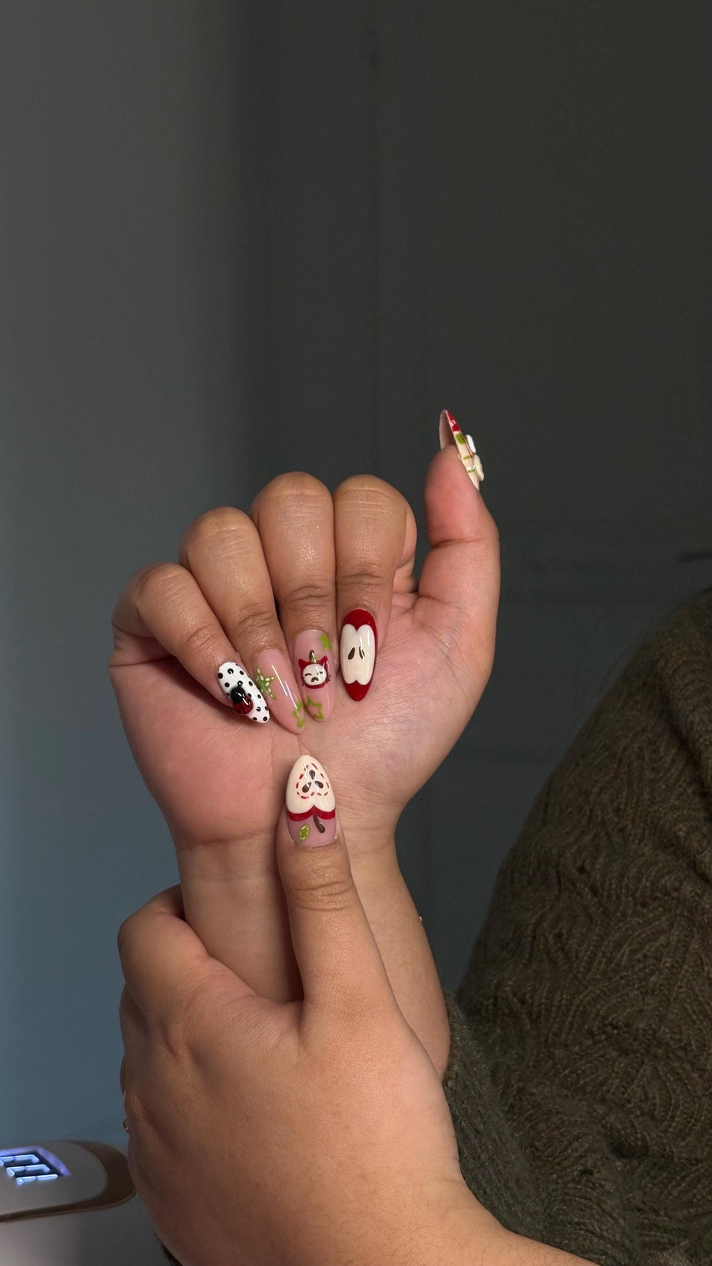 Press-on nail featuring apple, cat and button motifs in shades of red, green, and beige/ Faux ongles "press-on" avec motifs de pommes, chats et boutons, dans les tons rouge, vert et beige.